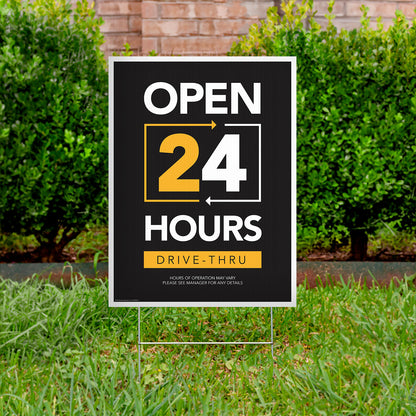 black, yellow, and white drive thru open 24 hours lawn sign in grass with bushes