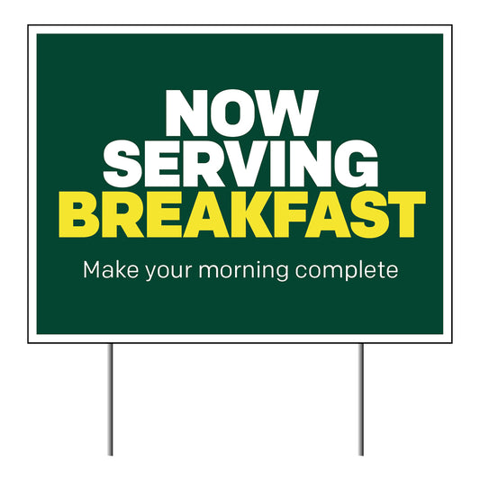 A green lawn sign with white and yellow text that reads 'NOW SERVING BREAKFAST Make your morning complete'.