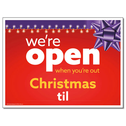 A red lawn sign with white and yellow text that reads 'we're open when you're out Christmas til' with a purple bow and Christmas lights depicted on the sign.